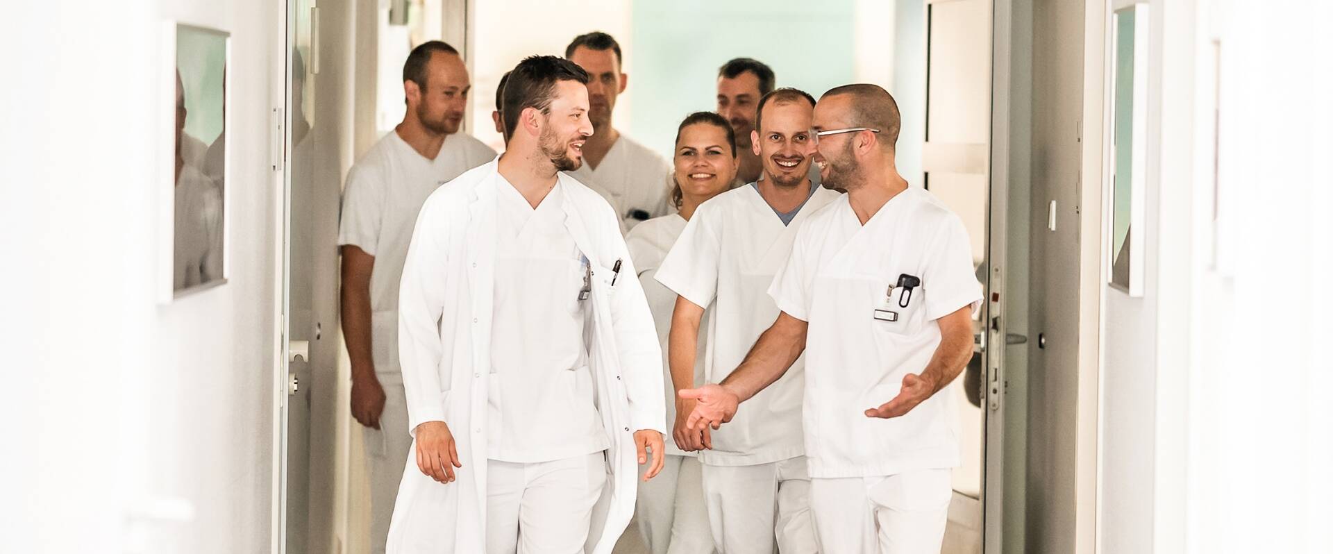 Eine Gruppe von Ärztinnen und Ärzten in weißer Arbeitskleidung gehen einen Flur entlang und unterhalten sich angeregt