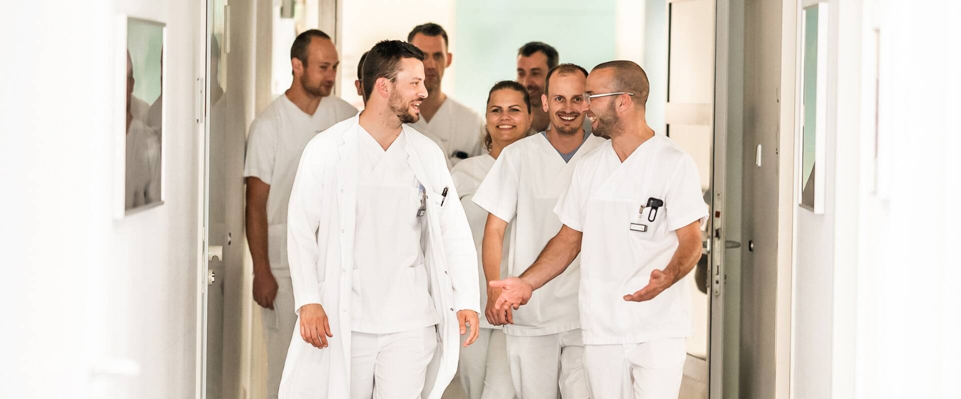 Eine Gruppe von Ärztinnen und Ärzten in weißer Arbeitskleidung gehen einen Flur entlang und unterhalten sich angeregt