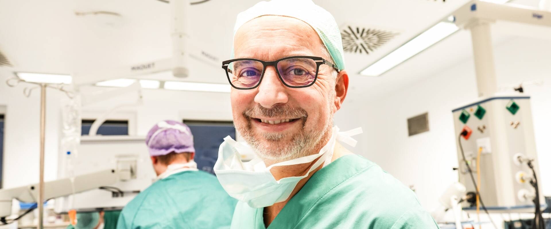 Freundlich lachender Arzt in OP-Montur mit herunterhängendem Mundschutz